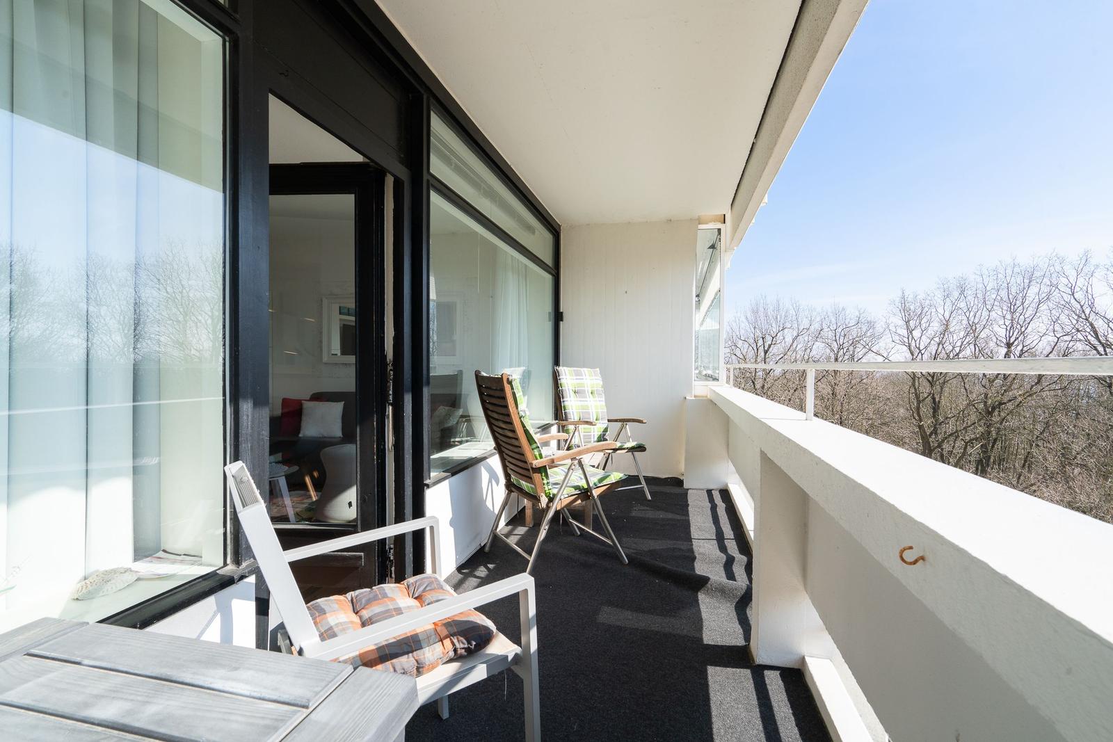 Terrace with chairs and table, view of trees. Glass facade leads into interior.