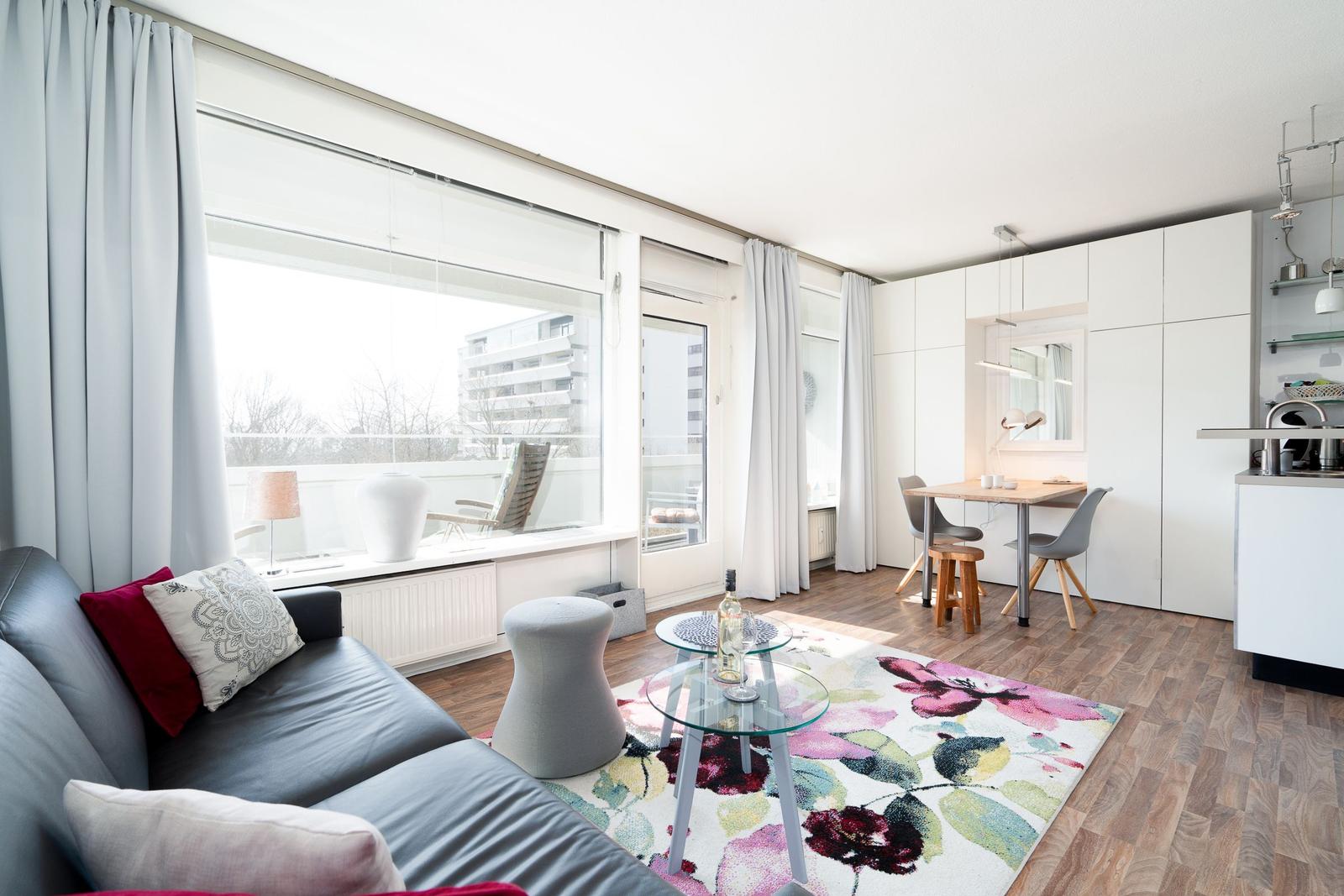 Living area with sofa, glass table, and floral rug. Dining area with table and chairs. Kitchen and balcony visible.