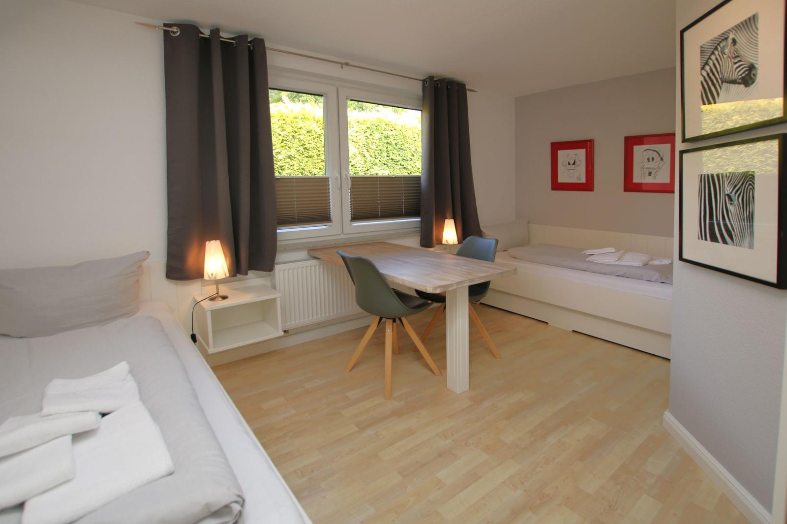 Zimmer mit zwei Betten, Tisch und Stühlen, Fenster mit Jalousien und Wandbildern.