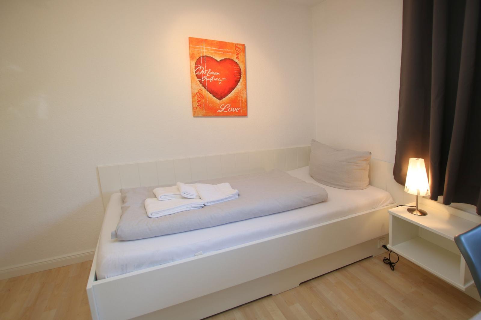 Ein Schlafzimmer mit weißem Bett, Wandbild und Nachttisch mit Lampe.