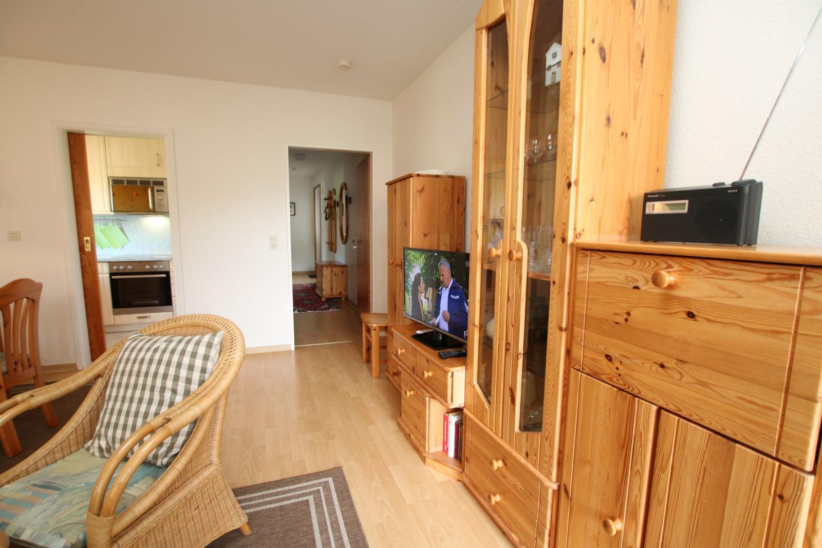 Wohnzimmer mit Holzmöbeln, Fernseher und Sitzgelegenheit. Küche und Flur sind sichtbar.