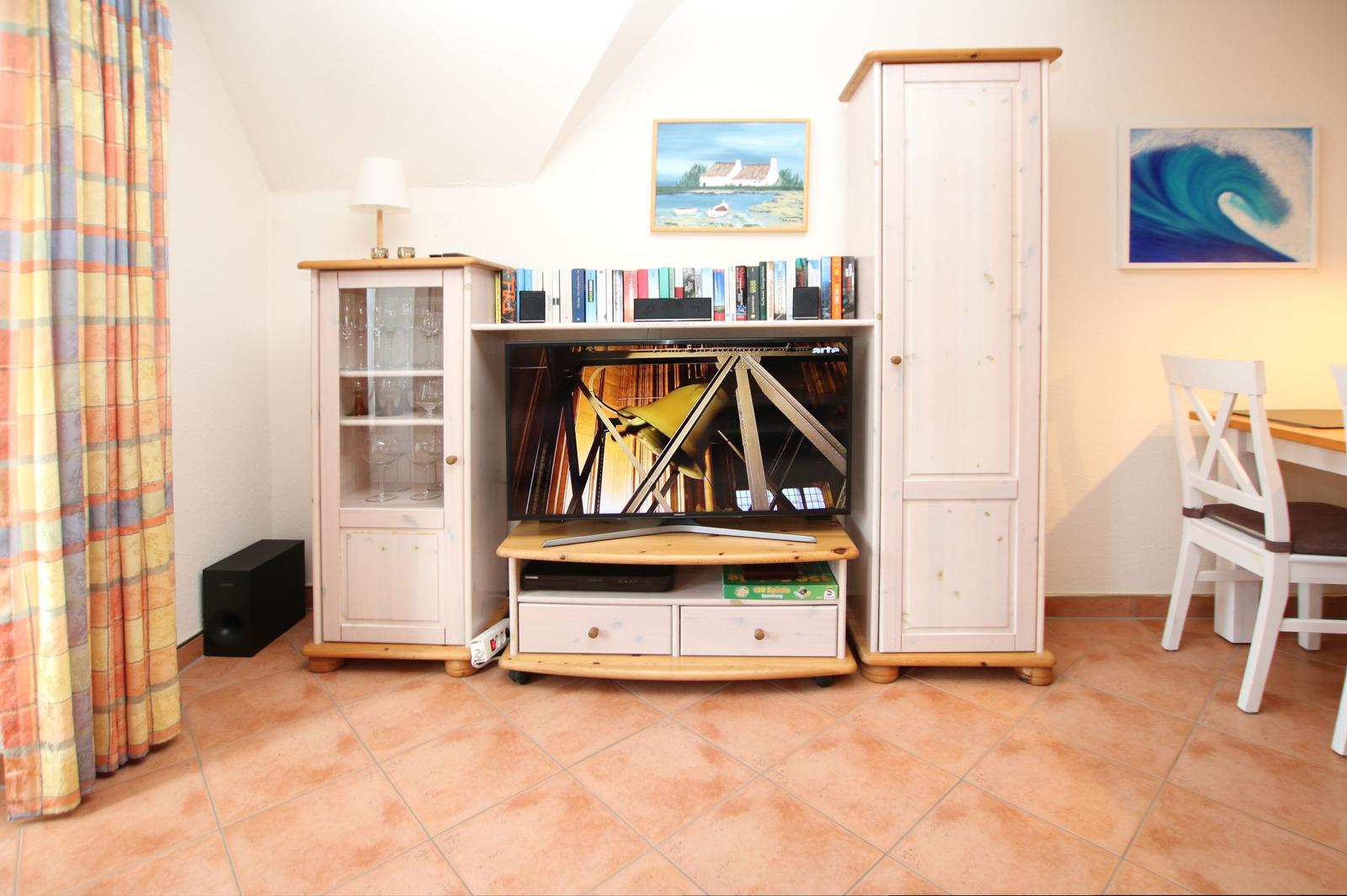 Living room with TV, bookshelf, and wardrobe. Terracotta floor and wall art.