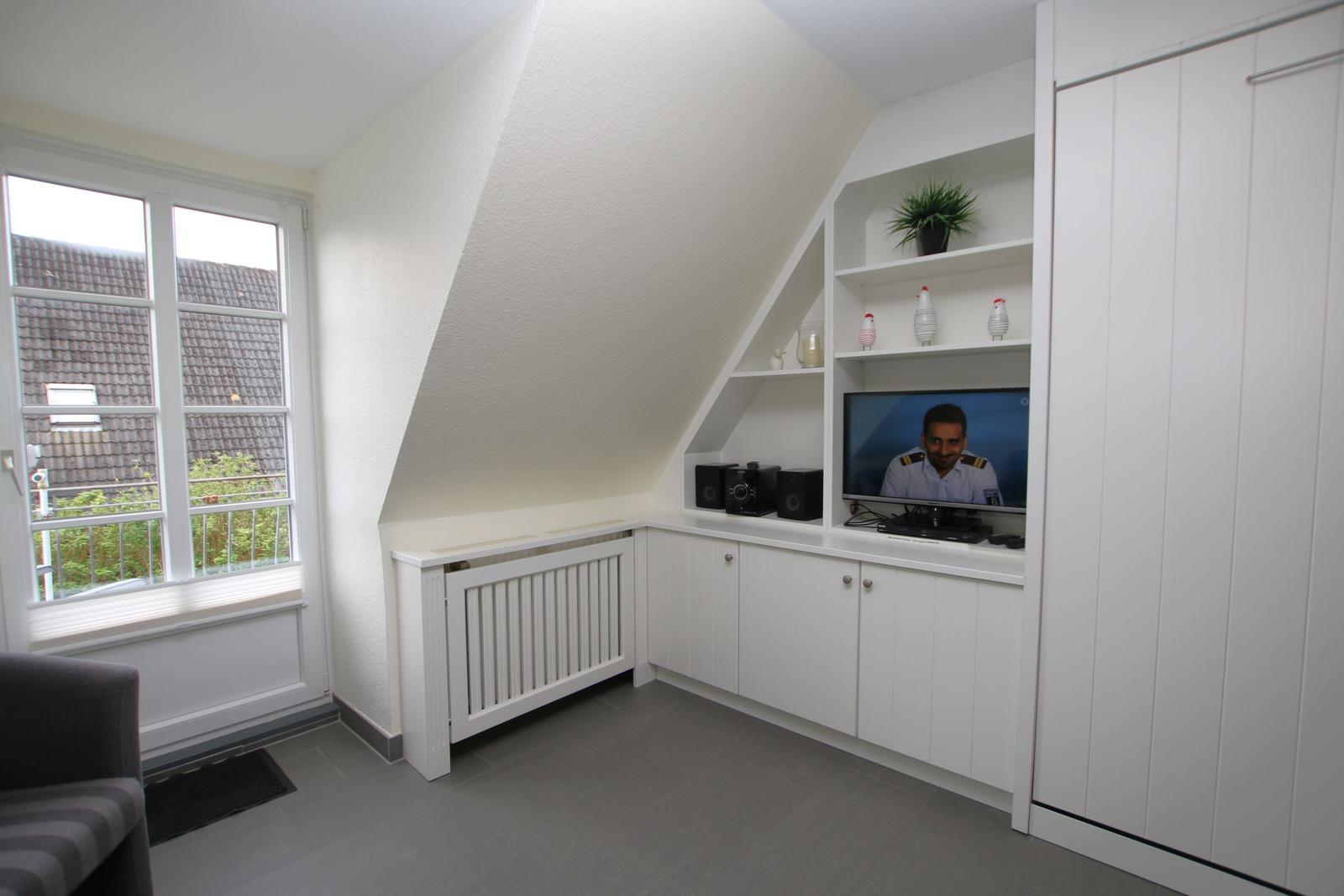 A room with sloped ceilings, window, TV, and white cabinets.