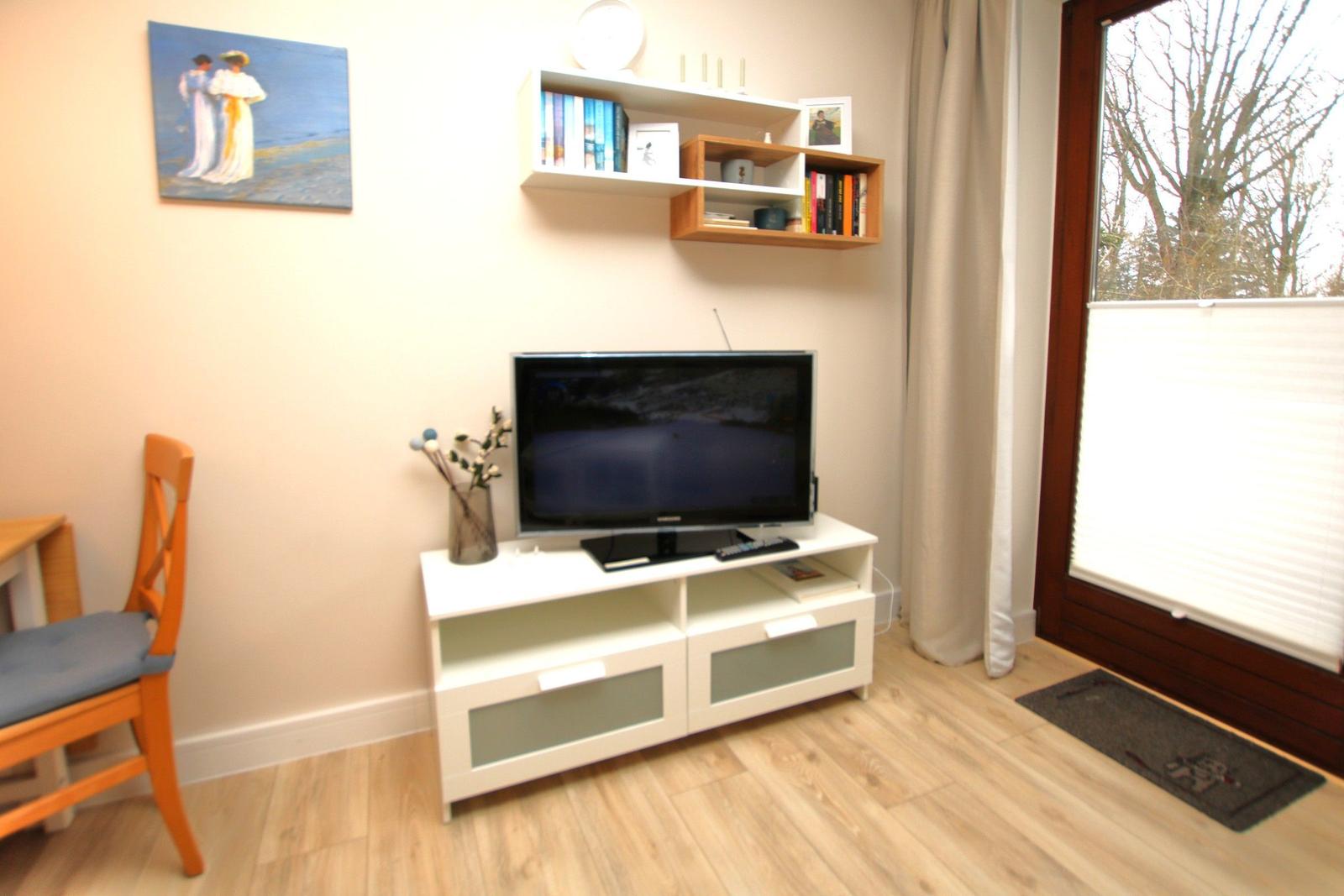 TV auf weißem Schrank, Wandregal mit Büchern, Fenster mit Jalousie und Holzboden.