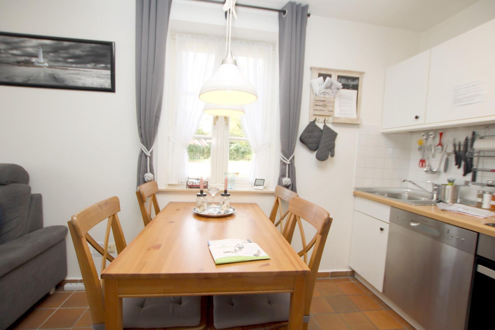 Essbereich mit Holztisch, Stühlen und Küchenzeile. Fenster mit Vorhängen und Wandbild.