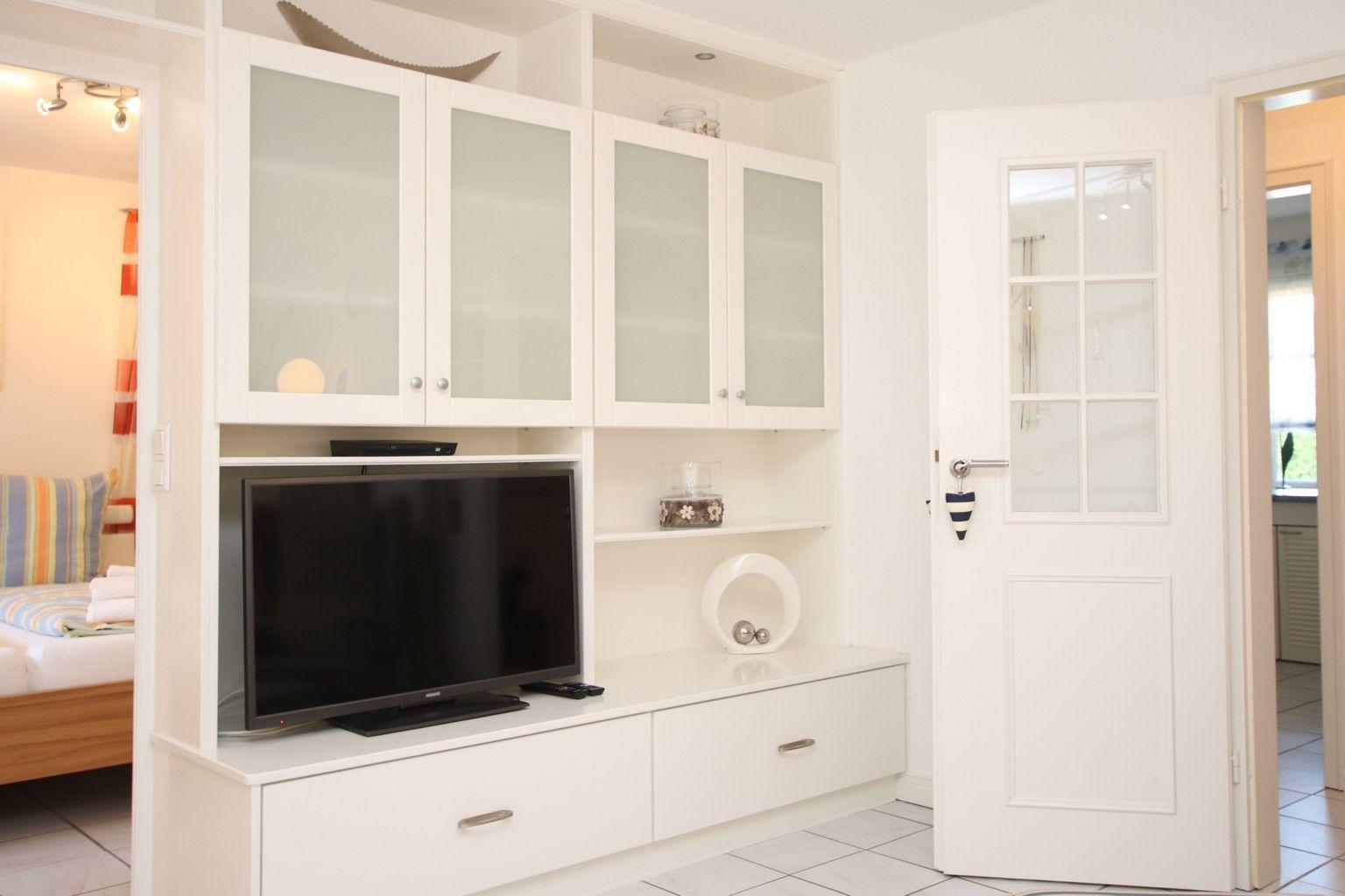 Weißer Schrank mit Fernseher und offenen Regalen. Tür mit Glasfenstern führt in einen anderen Raum.
