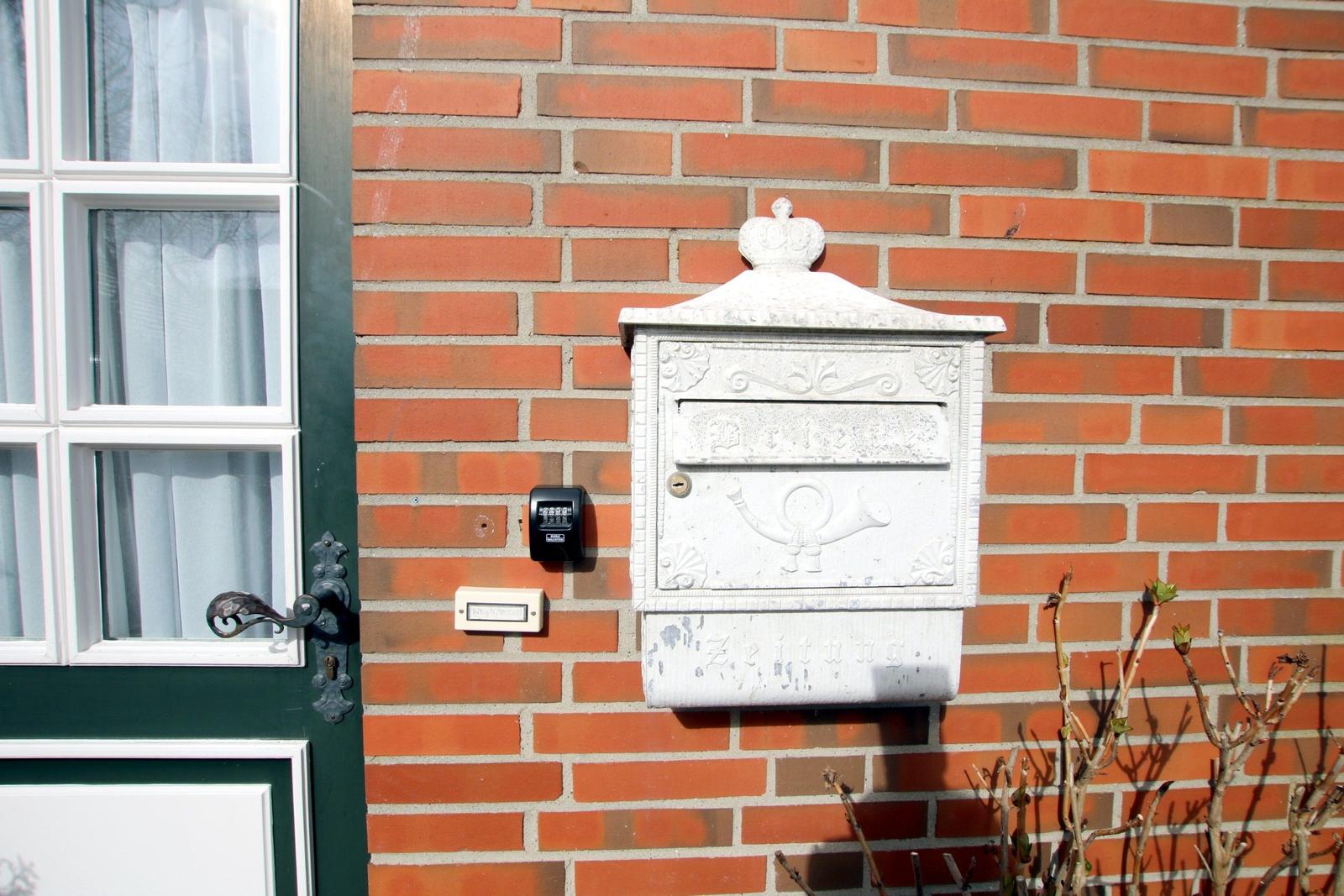 Weißer Briefkasten an roter Ziegelwand neben grüner Tür mit Fenster.