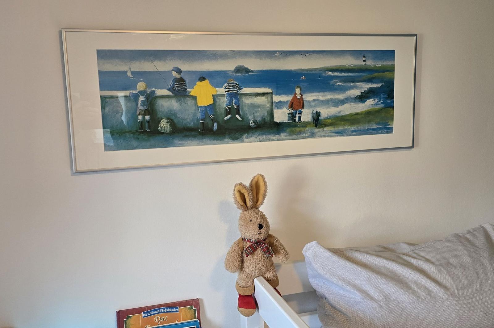 Wandbild mit Kindern am Meer über einem Bett mit Stoffhasen und Buch.