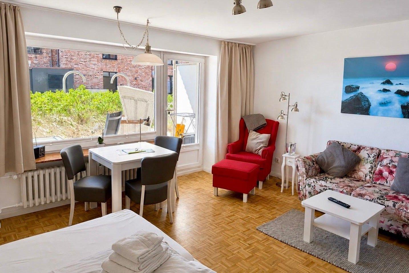 Living space with sleeping area, dining table, red armchair, and floral-patterned sofa.