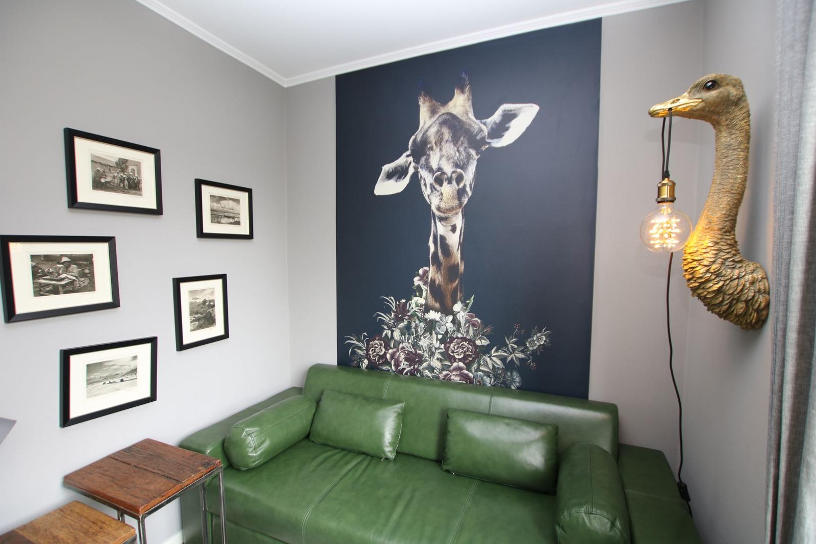 Ein Raum mit grünem Sofa, Wandbild einer Giraffe und einer Lampe in Form eines Straußens.