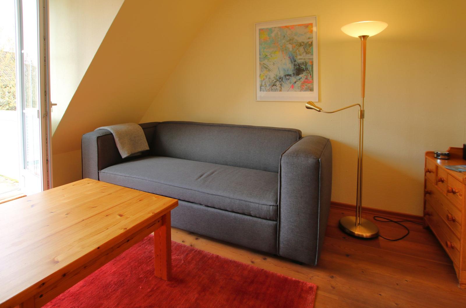 Ein Wohnzimmer mit grauem Sofa, Holztisch und Stehlampe. Wandbild und Holzfußboden sind sichtbar.