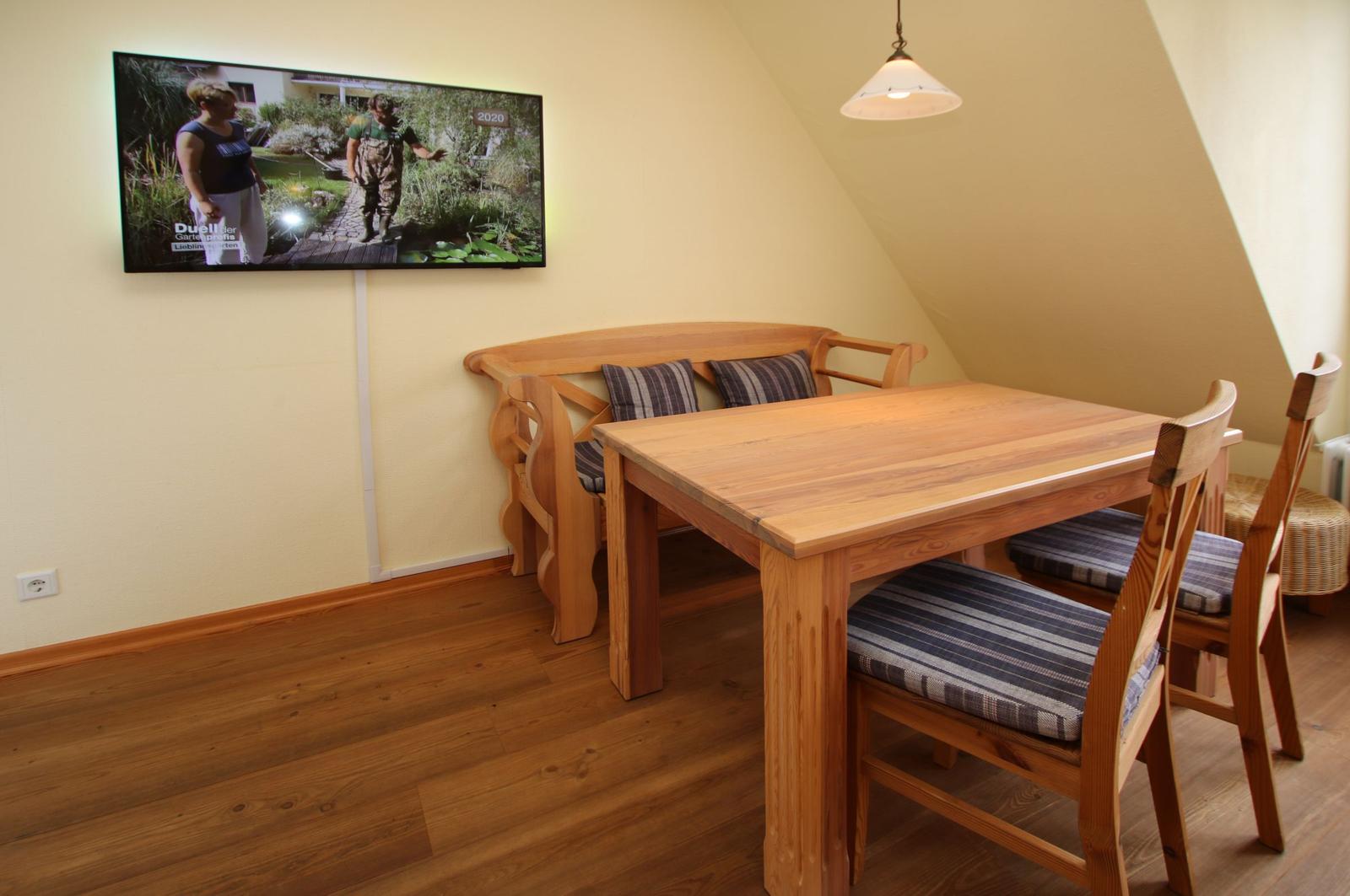 Esszimmer mit Holztisch, Stühlen und Sofa. Wandfernseher an der Wand.