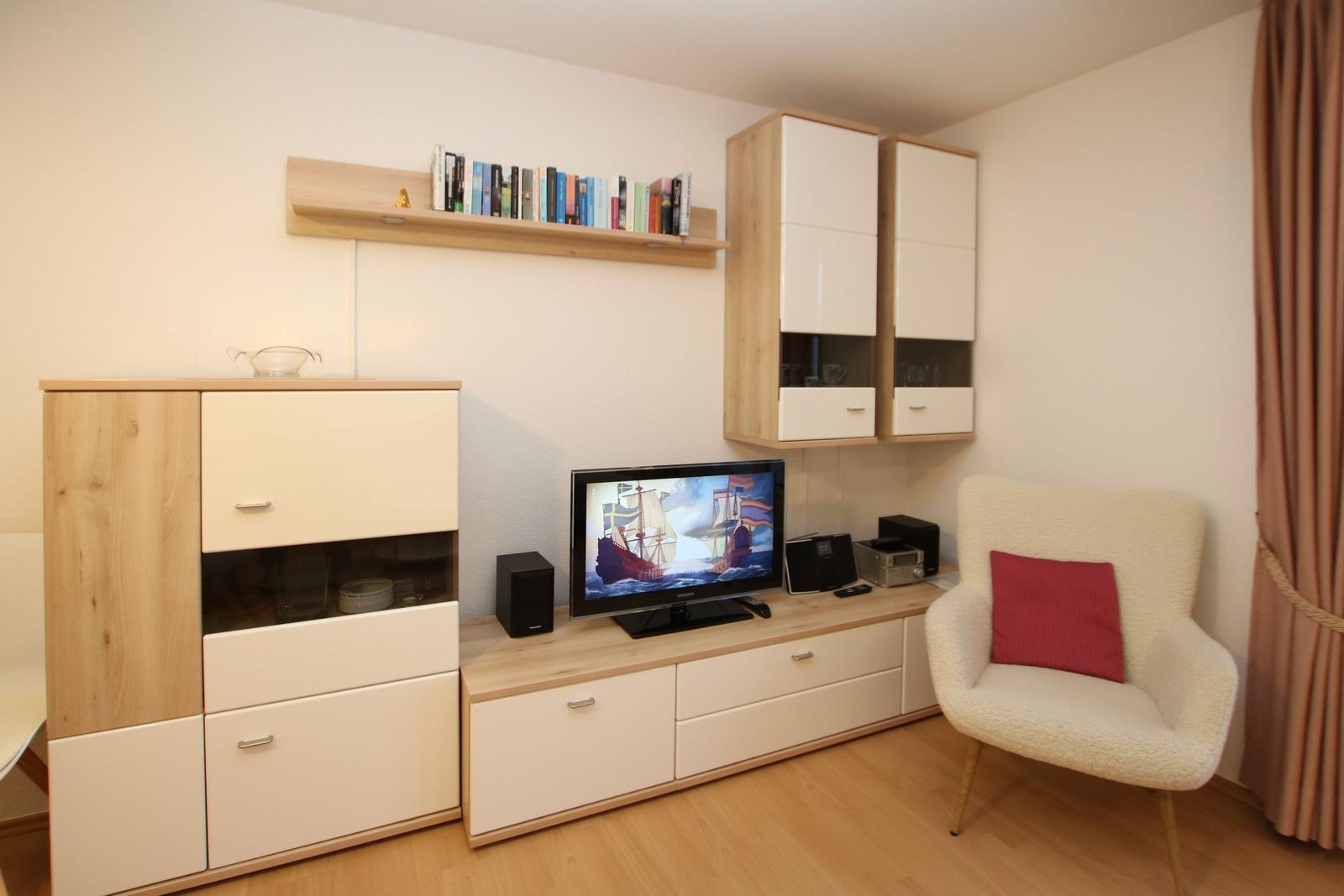 Ein Raum mit TV, Schränken und einem Sessel mit Kissen.