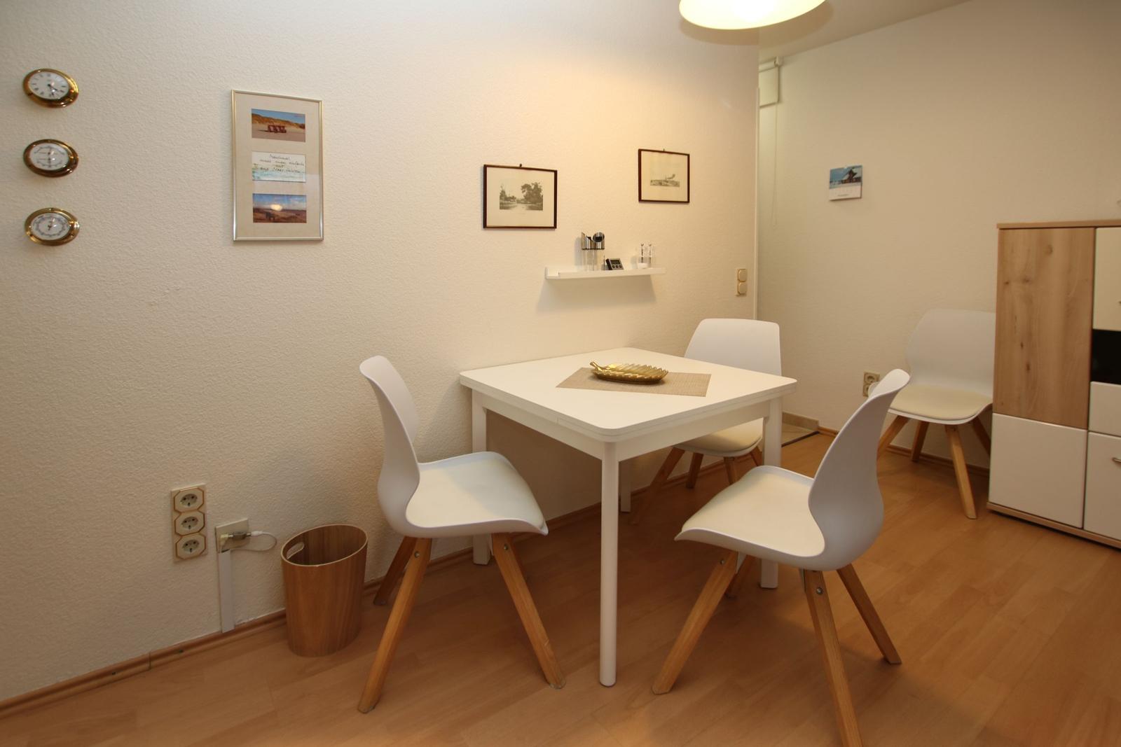 Esszimmer mit weißem Tisch, vier Stühlen und Wandbildern. Holzfußboden und Schrank im Hintergrund.