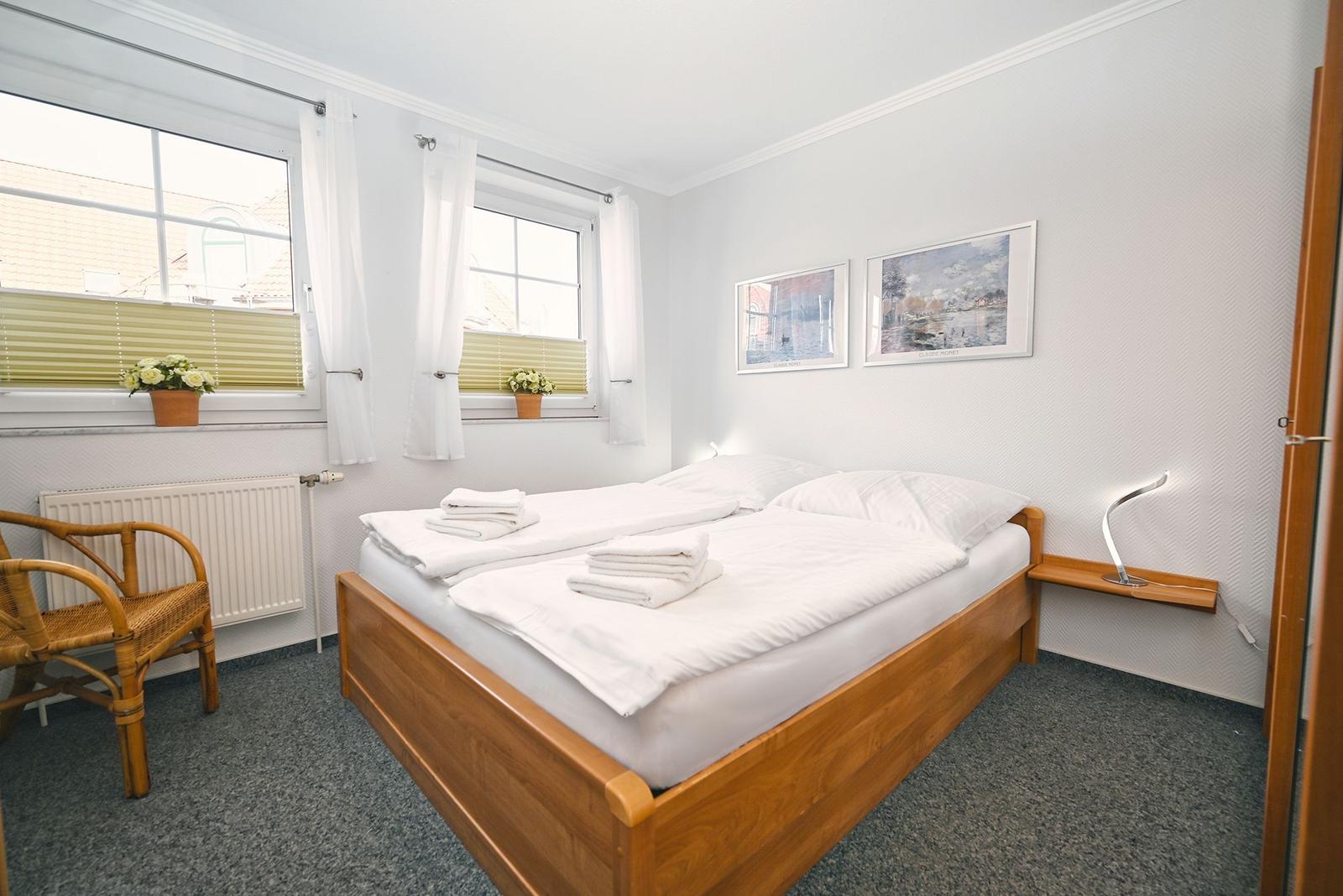 Schlafzimmer mit Doppelbett, zwei Fenstern und einem Stuhl.