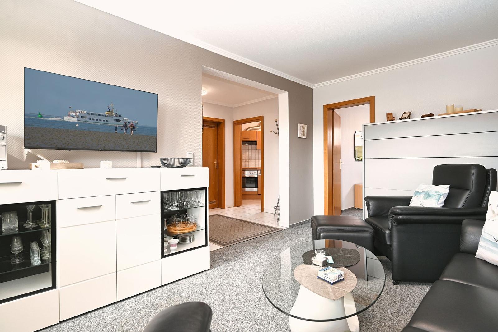 Wohnzimmer mit Fernseher, Couch und Glas-Tisch. Küche und Schlafzimmer sind sichtbar.