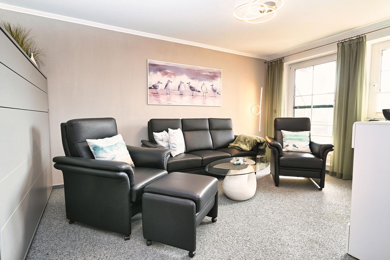 Wohnzimmer mit schwarzen Ledersofa und Sesseln, Glas-Tisch und Wandbild mit Möwen.