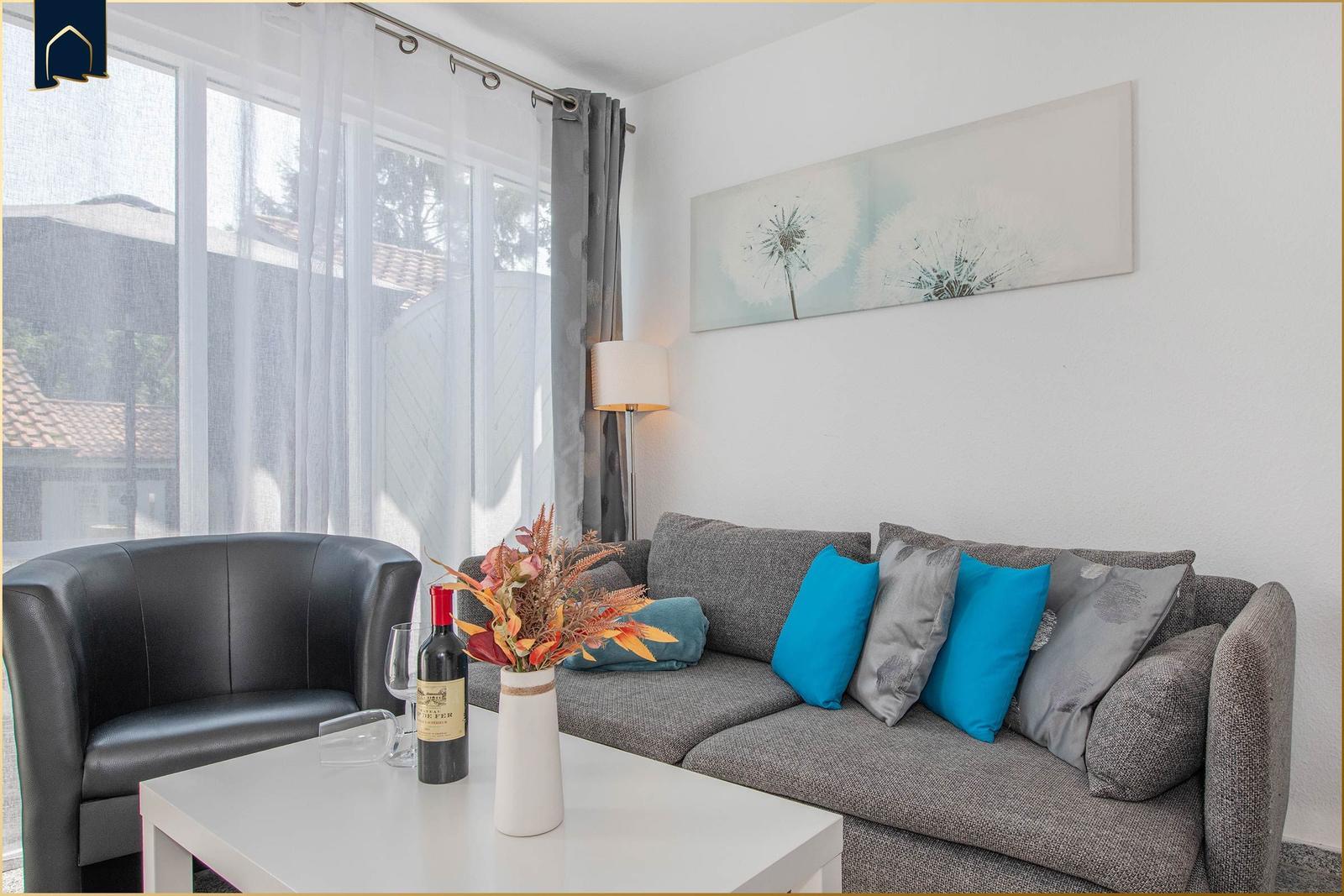 Ein Wohnzimmer mit grauem Sofa, blauen Kissen und einem schwarzen Sessel. An der Wand hängt ein Bild.