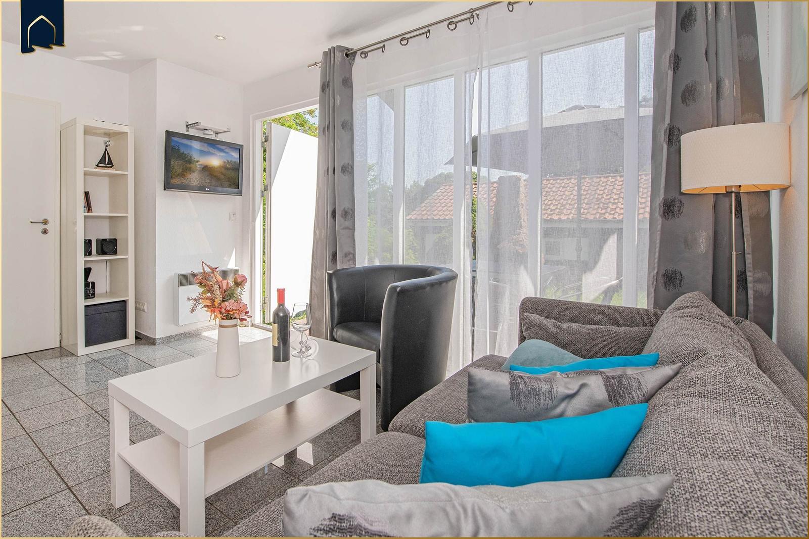 Wohnzimmer mit Sofa, Couchtisch und TV. Große Fenster mit Blick auf Außenbereich.