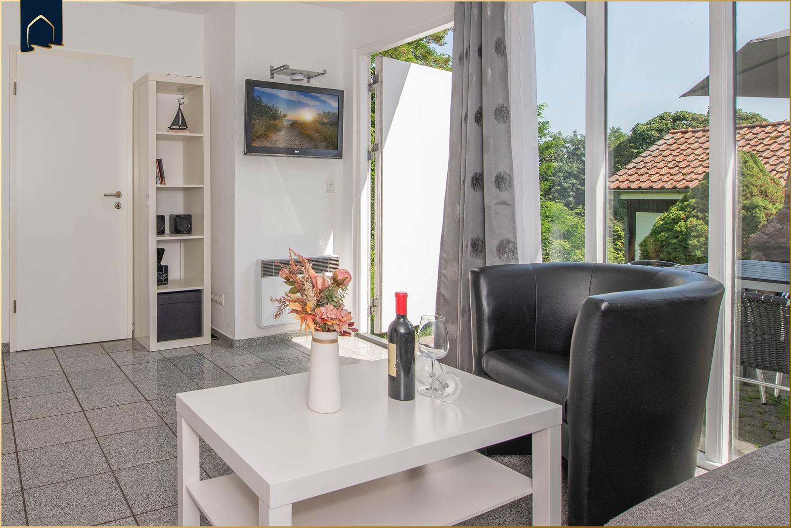 Wohnbereich mit schwarzer Couch, weißem Tisch und TV. Durchgehende Glasfront mit Blick auf Garten.