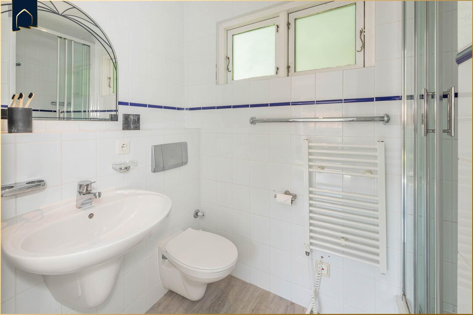 Badezimmer mit Waschbecken, Toilette und Dusche. Weiße Fliesen und Fenster.