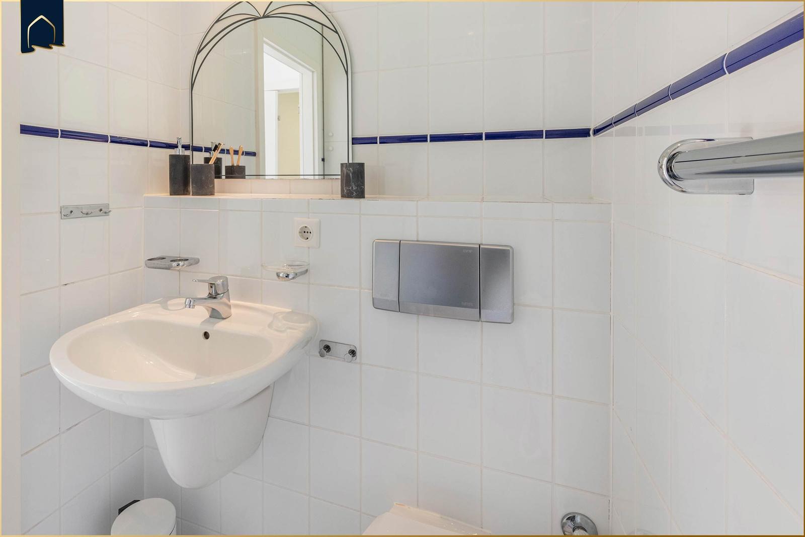Badezimmer mit Waschbecken, Spiegel und Toilette. Weiße Fliesen mit blauer Bordüre.