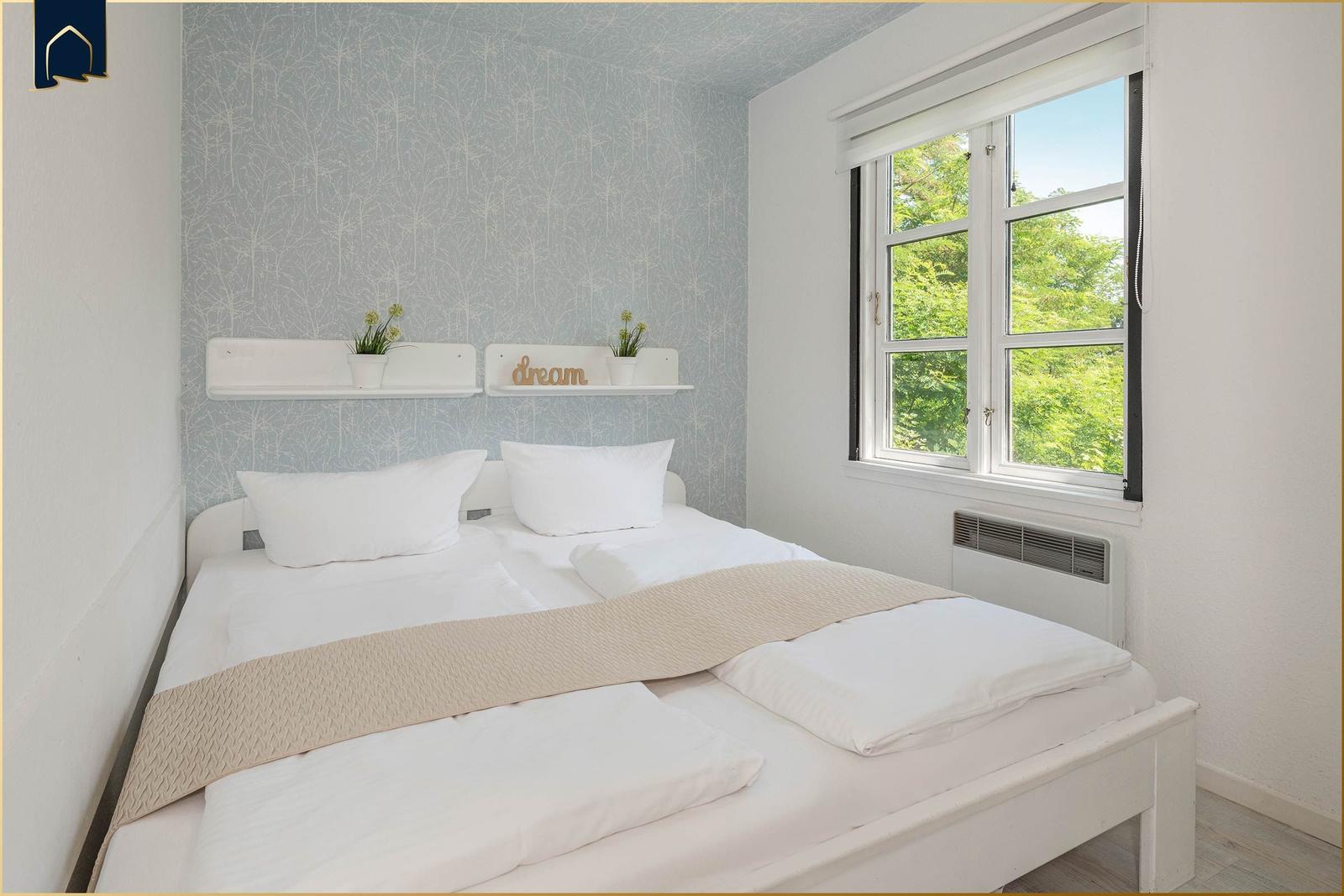 Schlafzimmer mit Doppelbett, Fenster mit Blick auf Bäume und Wand mit Tapete.