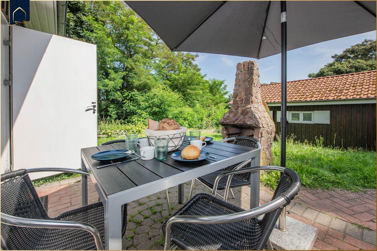 Terrasse mit Tisch, Stühlen und Sonnenschirm. Hintergrund: Steinofen und grüne Landschaft.
