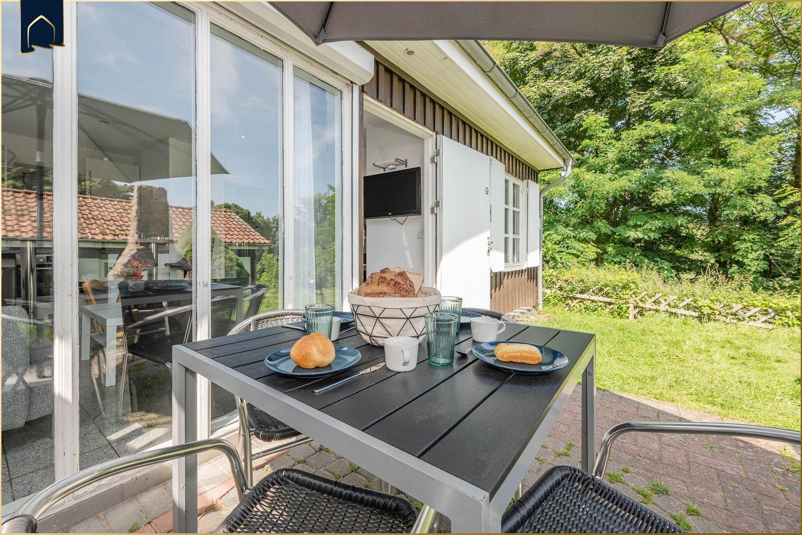 Terrasse mit Tisch, Stühlen und Brot. Hintergrund: Grünfläche und Haus.