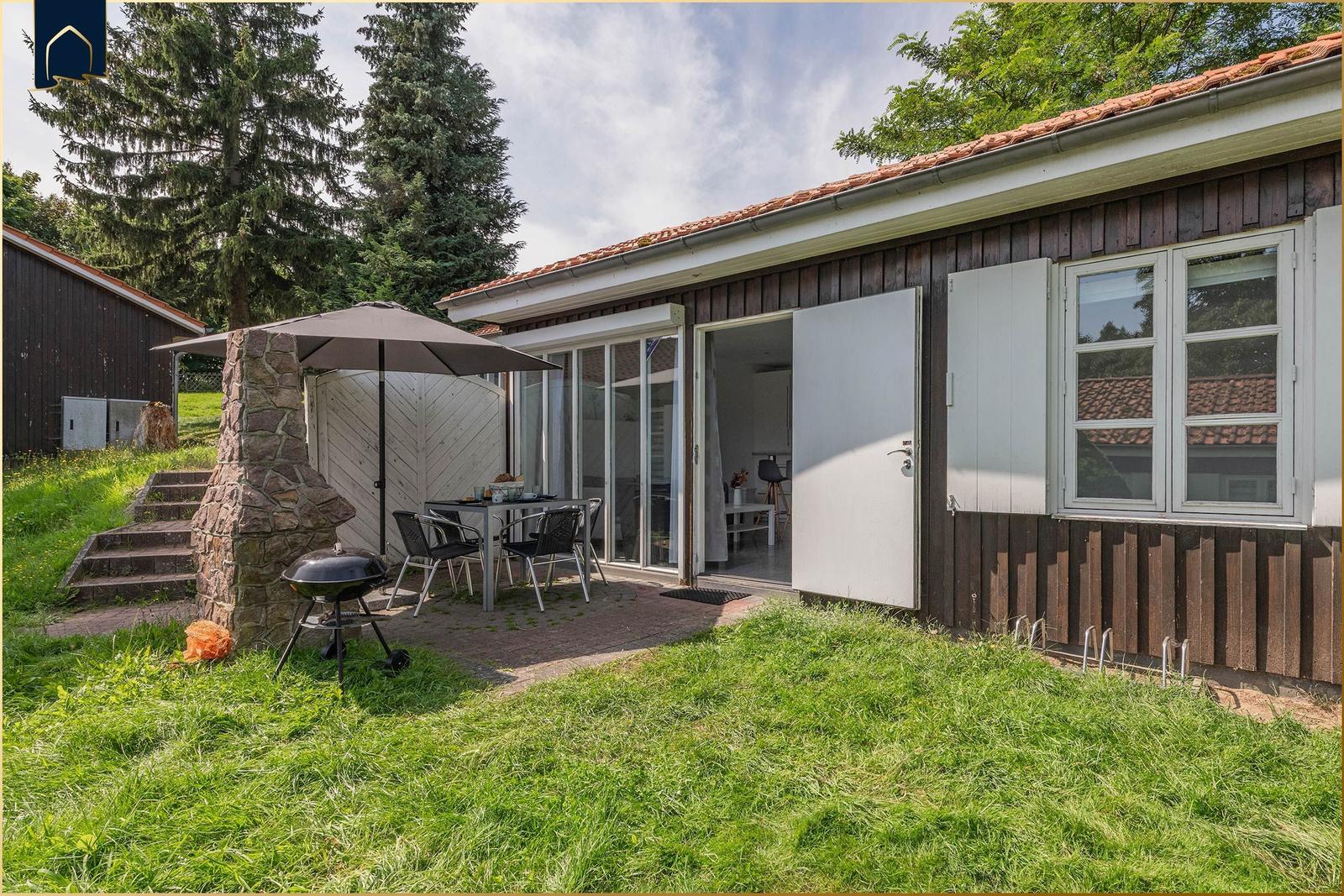Haus mit Terrasse, Grill und Sitzgruppe im Freien. Glasfront öffnet sich ins Innere.