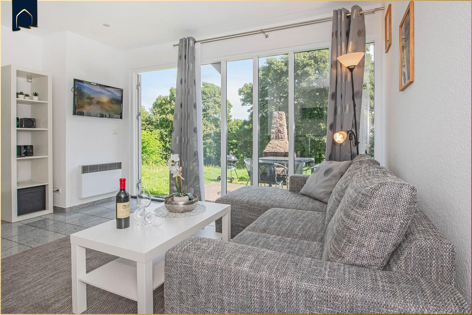 Wohnzimmer mit Sofa, Couchtisch und Fernseher. Durchführung zur Terrasse mit Blick auf Grünflächen.