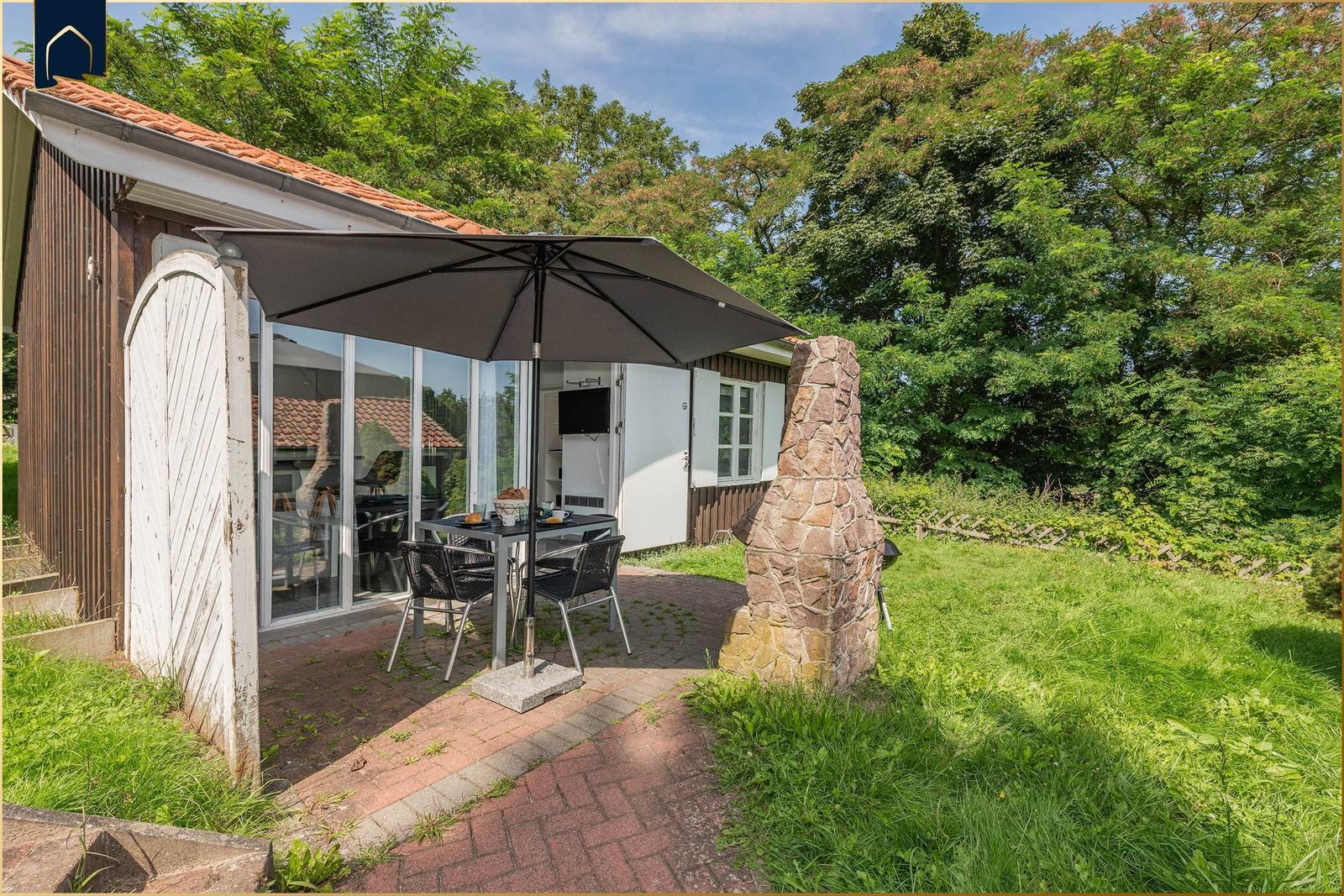 Terrasse mit Tisch, Stühlen und Sonnenschirm vor einem Haus mit Glasfront.