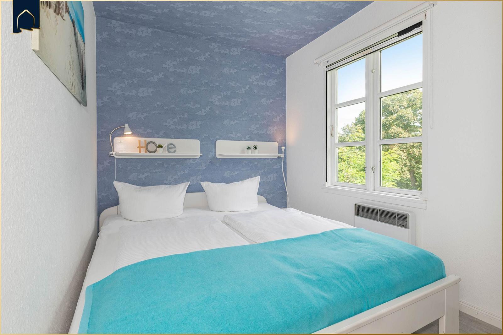 Schlafzimmer mit Bett, blauer Tapete und Fenster mit Blick auf Bäume.