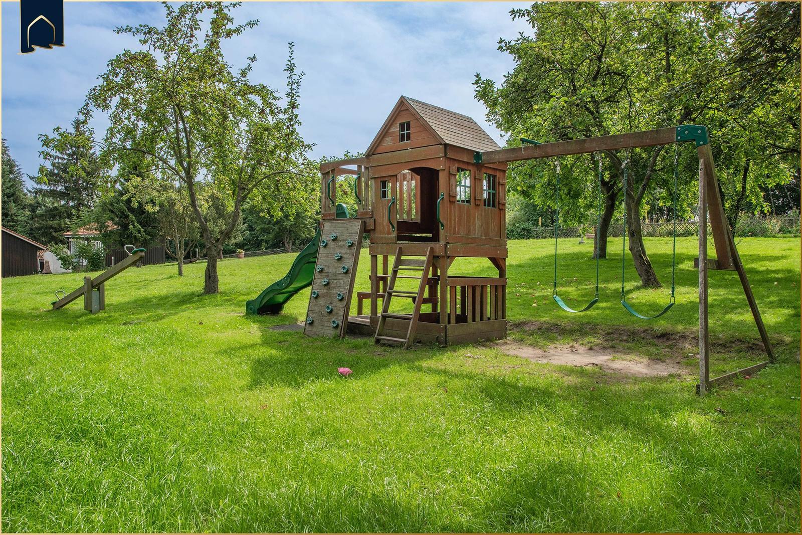 Großer Garten mit Holzspielhaus, Rutsche und Schaukel unter Bäumen.
