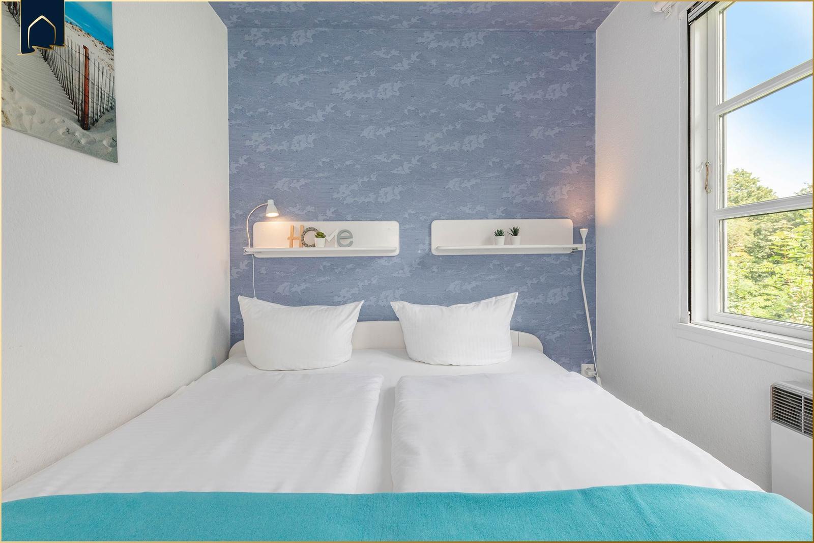 Schlafzimmer mit Doppelbett, blauer Tapete und Fenster mit Blick auf Bäume.