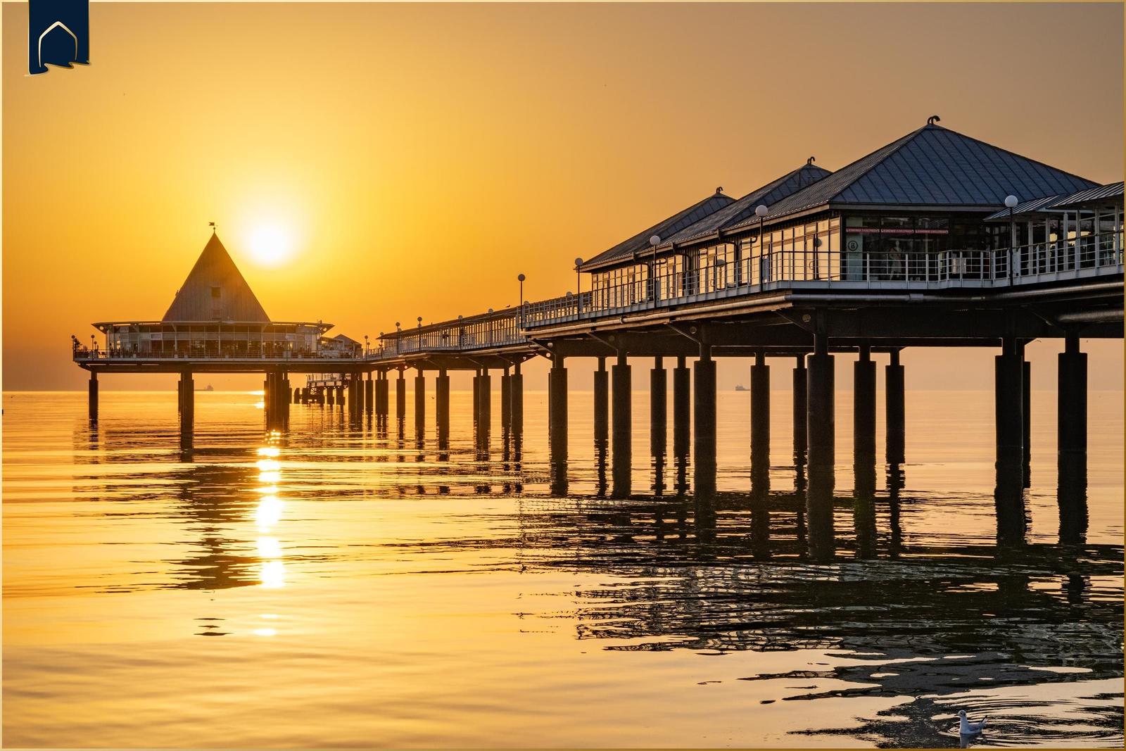 Sonnenuntergang über einem Steg mit Pavillon und Reflexion im Wasser.