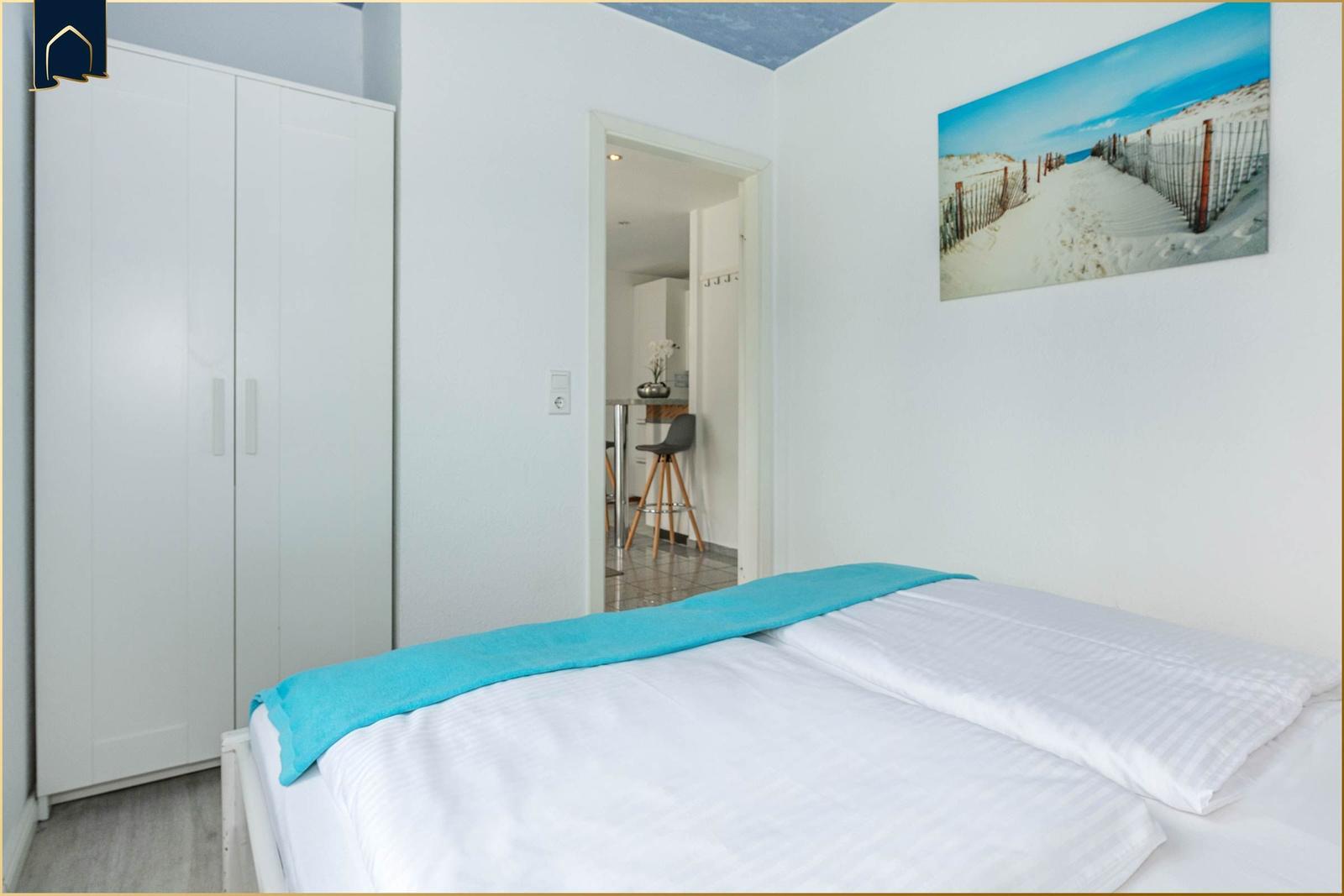 Schlafzimmer mit Doppelbett, weißer Kleiderschrank und Strandbild an der Wand.