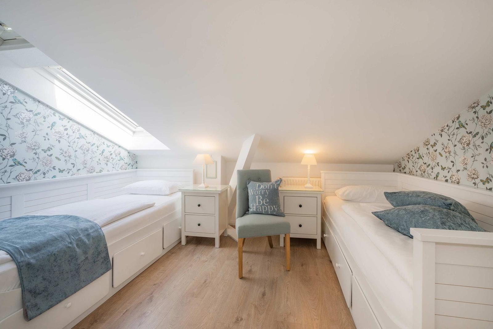 Zimmer mit zwei Einzelbetten, Nachttischen und Stuhl unter Dachschräge mit Blumenwand.