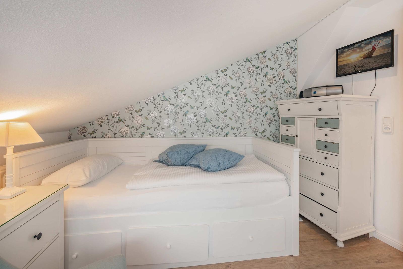 Schlafzimmer mit Doppelbett, Schrank und Wandbild. Decke geneigt, Tapete mit Blumenmuster.