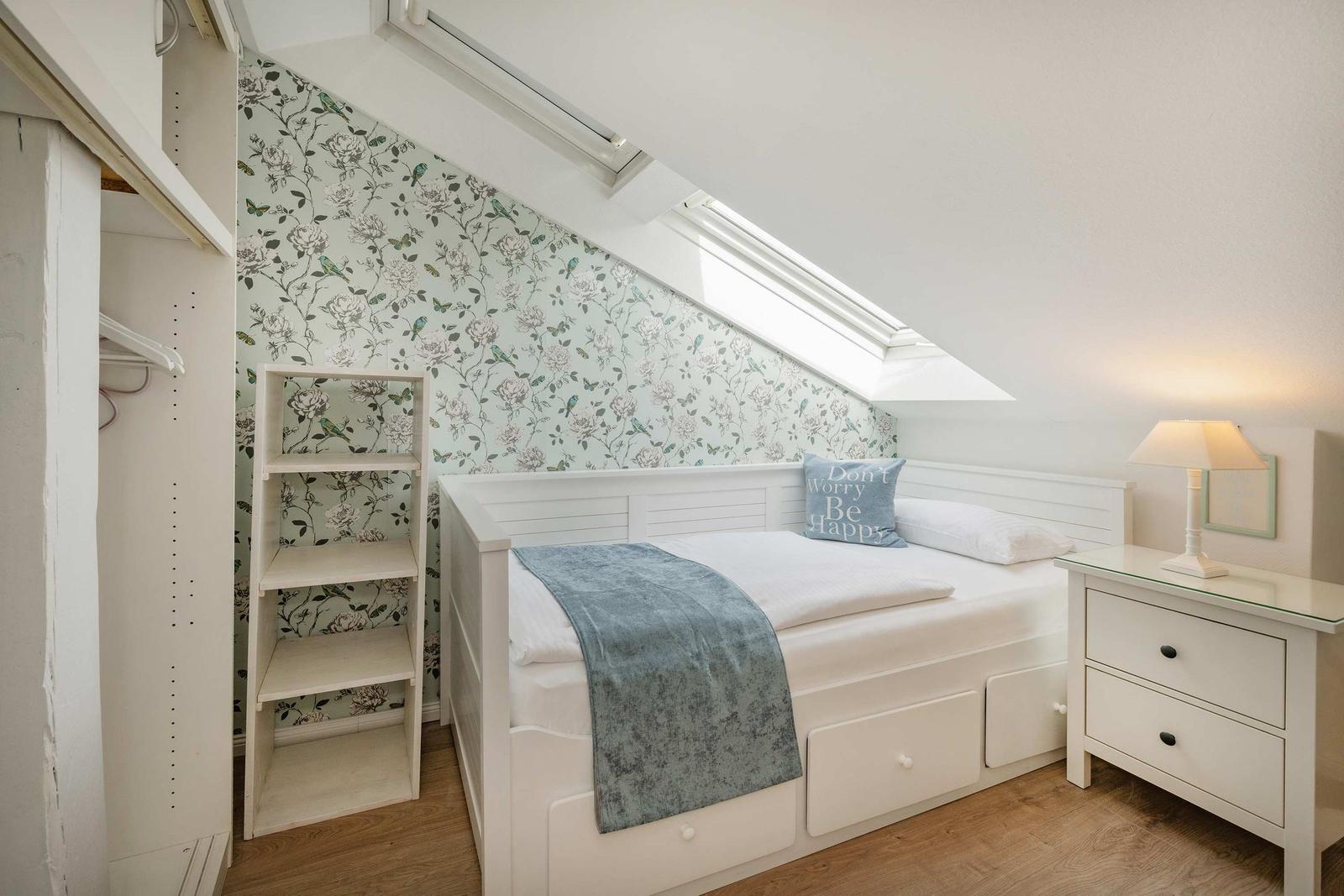 Schlafzimmer mit Dachschrägen, weißen Möbeln und floraler Tapete an der Wand.