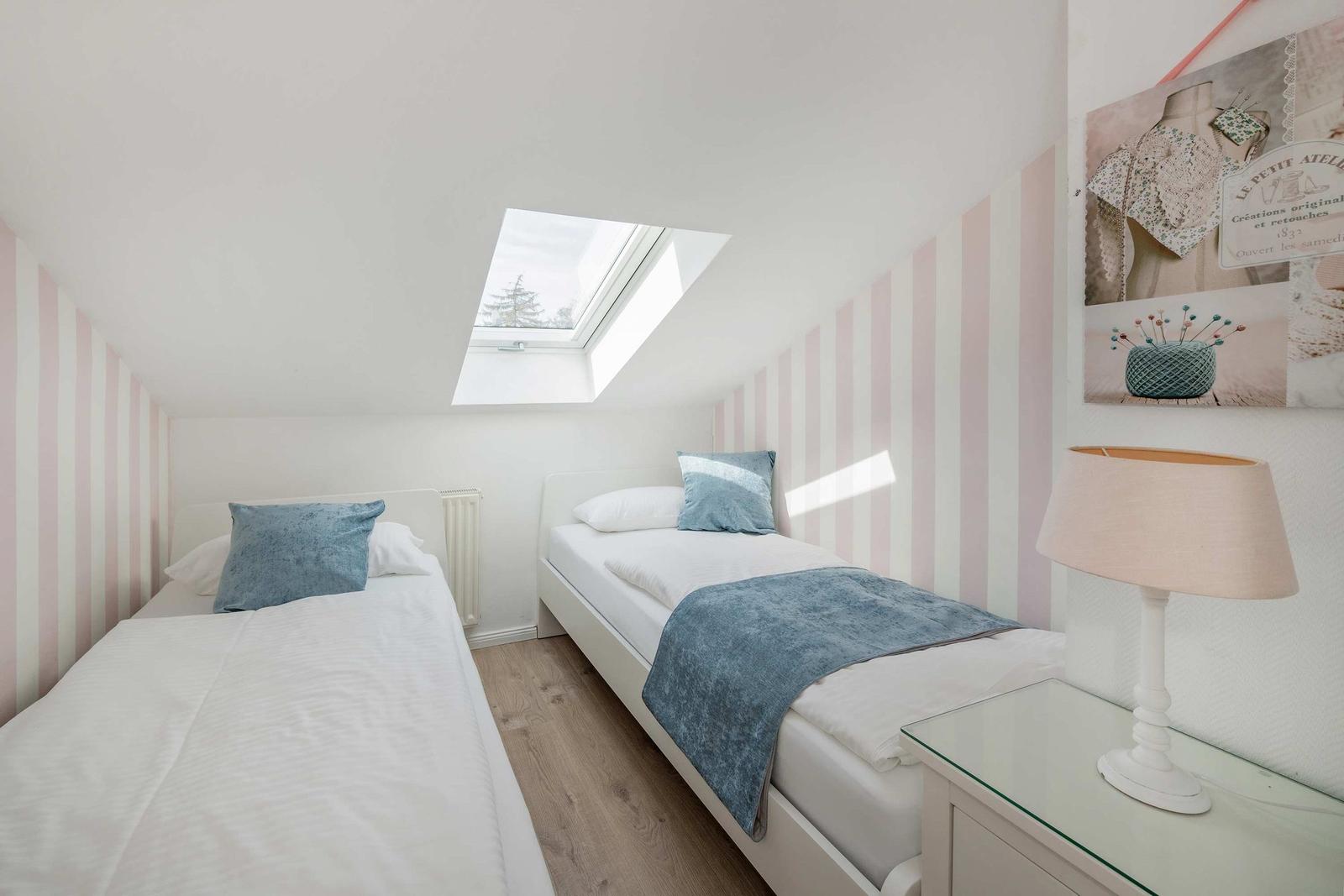 Zwei Einzelbetten unter Dachschrägen mit Fenster, rosa gestreifte Wände und Nachttisch mit Lampe.
