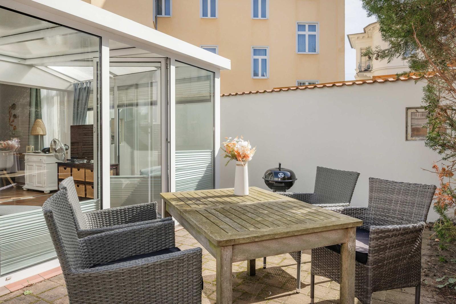 Terrasse mit Tisch, Stühlen und Grill. Glasveranda und Hauswand im Hintergrund.
