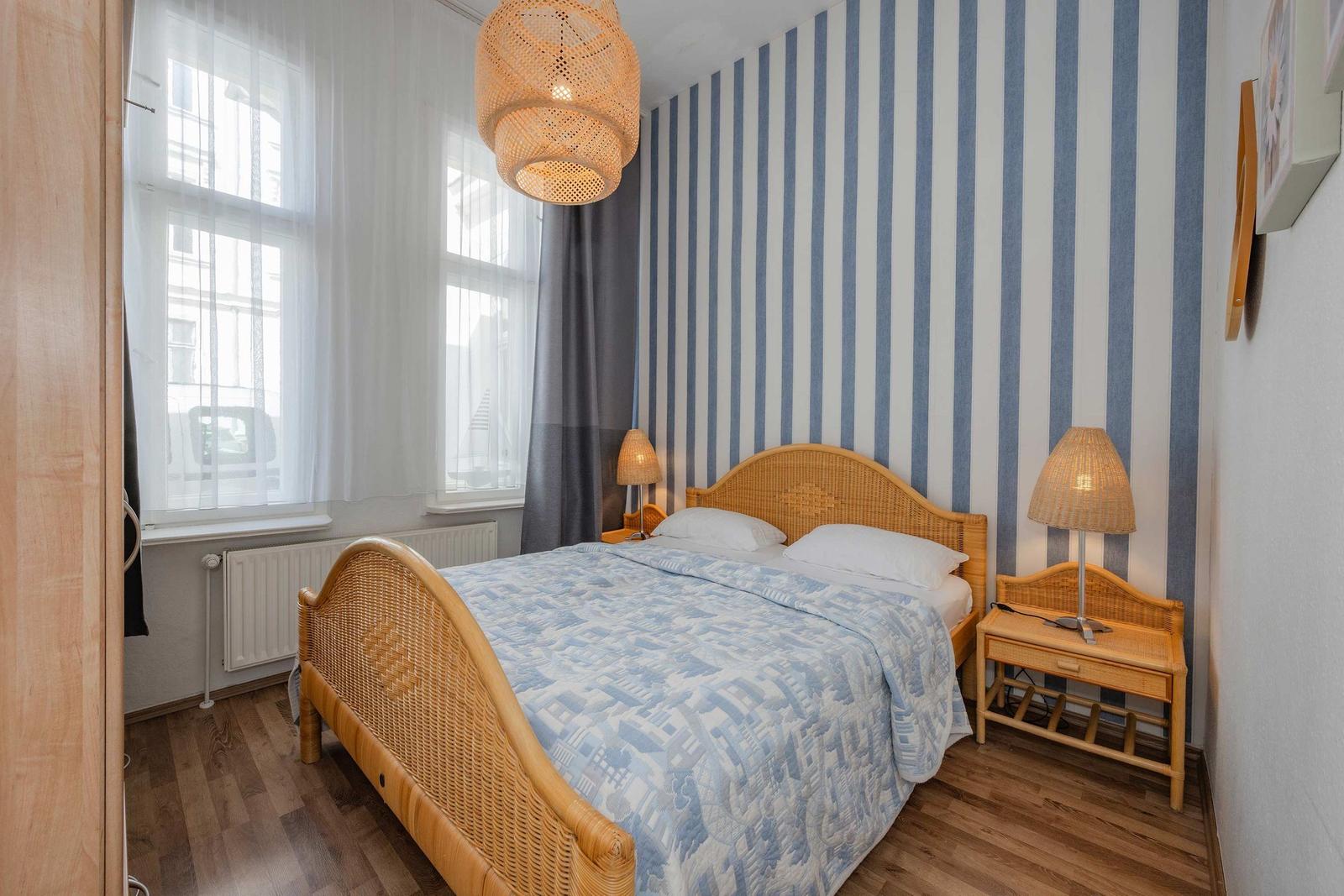 Schlafzimmer mit Bett, Nachttischen und Streifenwand. Fenster mit Gardinen.