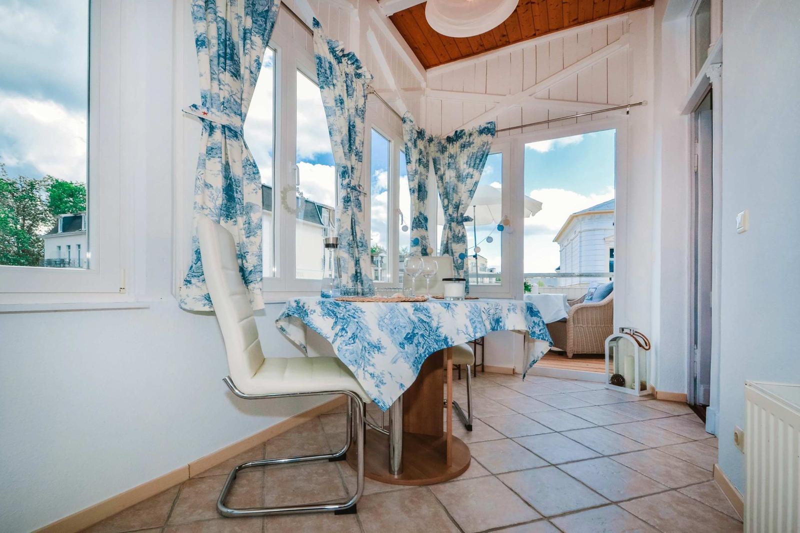 Dining table with chairs and blue-white tablecloth in front of windows with balcony view.