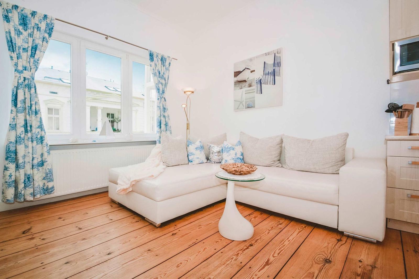 Living room with white sofa, wooden floor, and blue curtains. View of buildings through window.