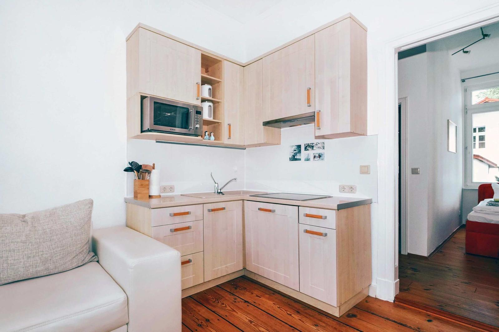 Kitchen with wooden cabinets and microwave. Living area with sofa. Wooden floor and white walls.