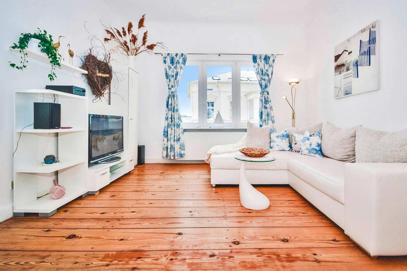 Living room with white sofa, wooden floor, and TV unit. Windows with blue curtains.