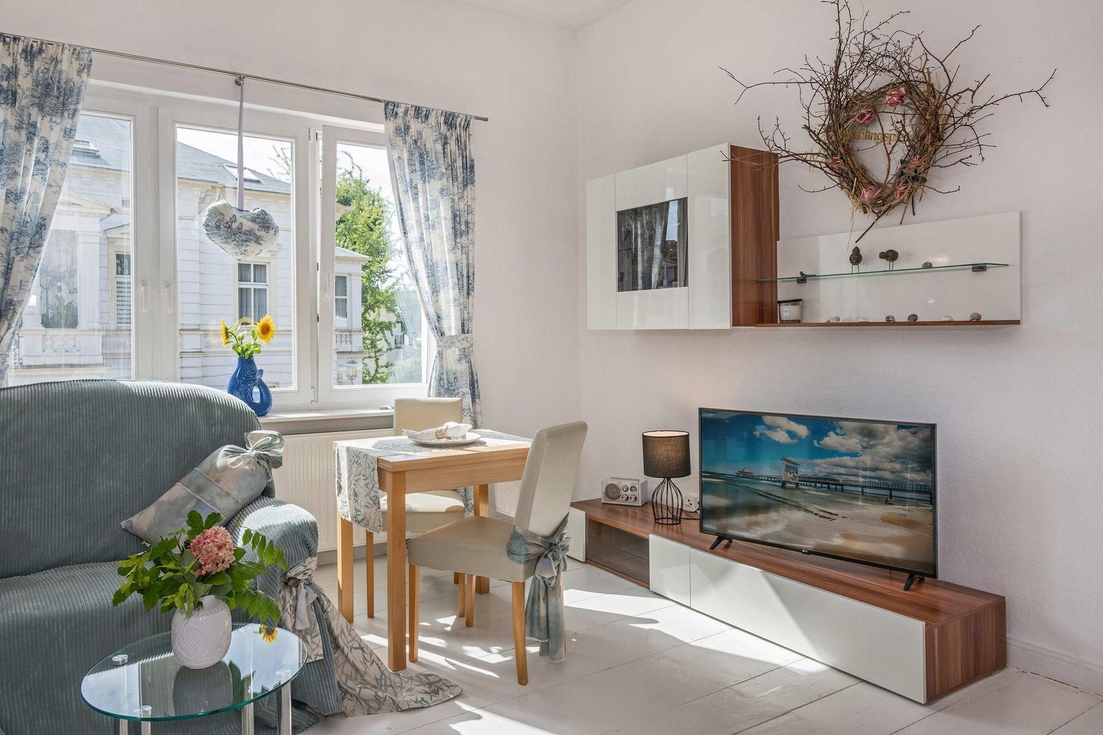 Wohnzimmer mit Sofa, Esstisch und Fernseher. Fenster mit Vorhängen und Blumen.