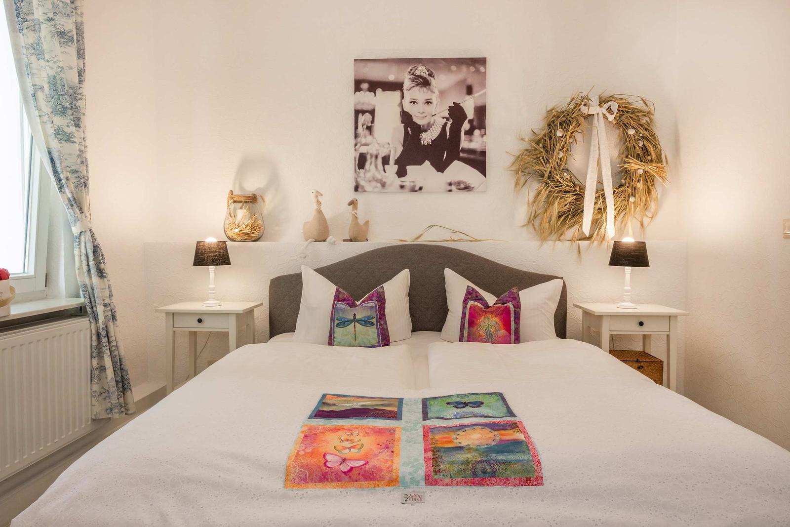 Schlafzimmer mit Bett, Nachttischen und Wandbild. Vorhang und Kissen mit Mustern.