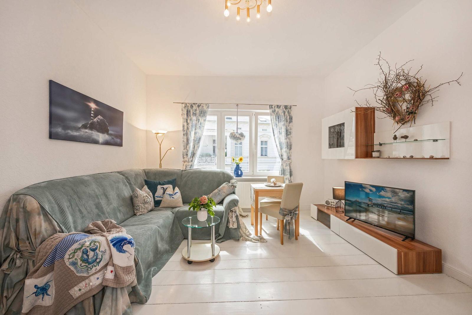 Wohnzimmer mit Sofa, Fernseher und Essplatz. Fenster mit Vorhängen. Wandbild und Deckenleuchte.